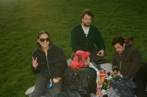 Four people enjoy a sunny picnic on a grassy field, sharing snacks and drinks in a relaxed, outdoor setting with casual attire.