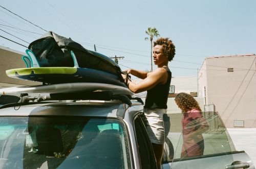 Two people by a car with surfboards on top. One person is loading boards while the other is next to the open car door. Sunny day.