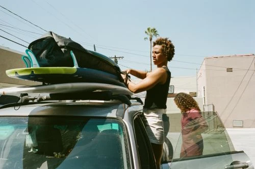 Two people by a car with surfboards on top. One person is loading boards while the other is next to the open car door. Sunny day.
