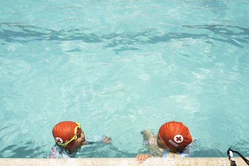 Two children in orange swimming caps with red crosses in an outdoor swimming pool during daytime, viewed from above the water.