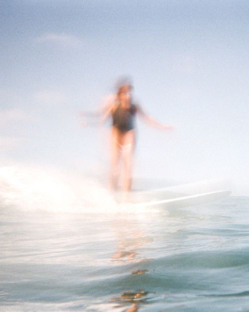 Blurred view of a person in a dark swimsuit surfing on a white surfboard in the ocean on a bright day with a blue sky.