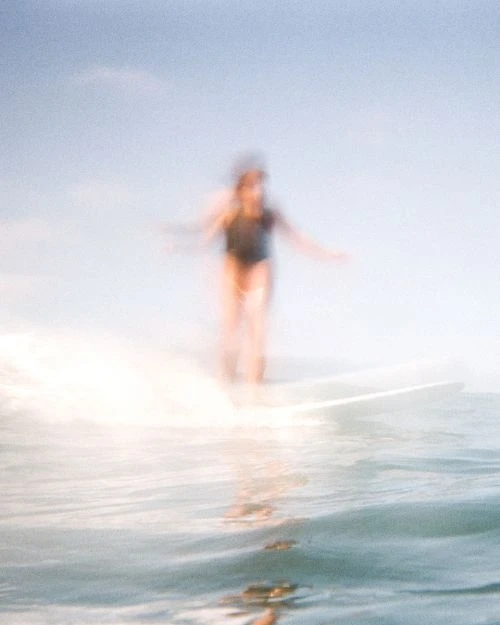 Blurred view of a person in a dark swimsuit surfing on a white surfboard in the ocean on a bright day with a blue sky.