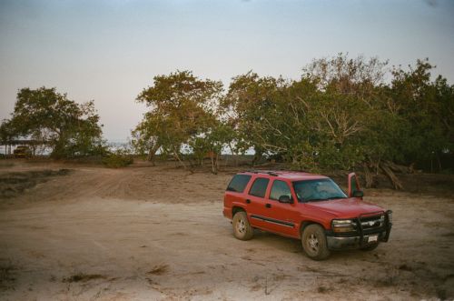 Red SUV parked on sandy shore with trees in the background under soft sky. Vehicle has an open door. Natural setting. 