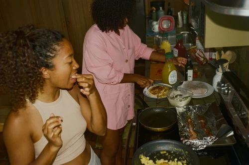 Two women in a home kitchen preparing food at the stove; one is tasting a snack while the other cooks breakfast or brunch.