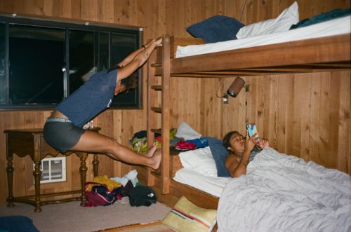 Two people in a cabin room. One woman stretches on bunk bed ladder, while another relaxes in the lower bunk bed, using a smartphone.
