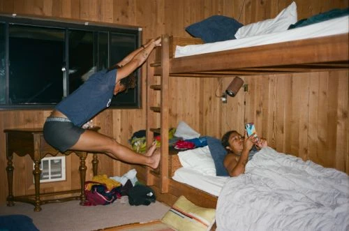 Two people in a cabin room. One woman stretches on bunk bed ladder, while another relaxes in the lower bunk bed, using a smartphone.