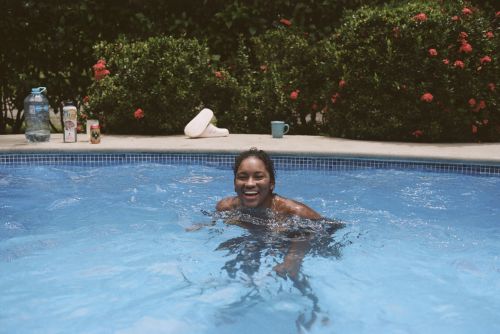 Smiling woman swimming in a blue tiled outdoor pool surrounded by green foliage and red flowers on a sunny day.