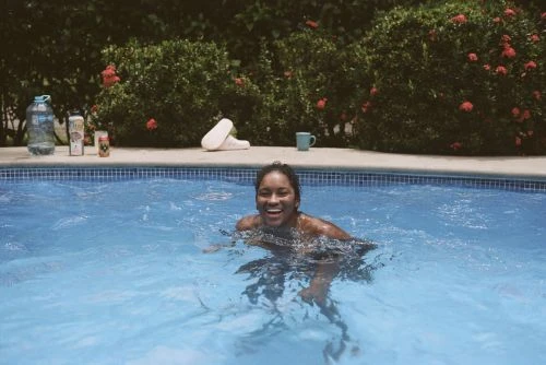 Smiling woman swimming in a blue tiled outdoor pool surrounded by green foliage and red flowers on a sunny day.