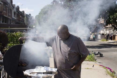 An African-American man stands outdoors on a sunny day tending a smoking barbecue grill in a residential street setting with houses.