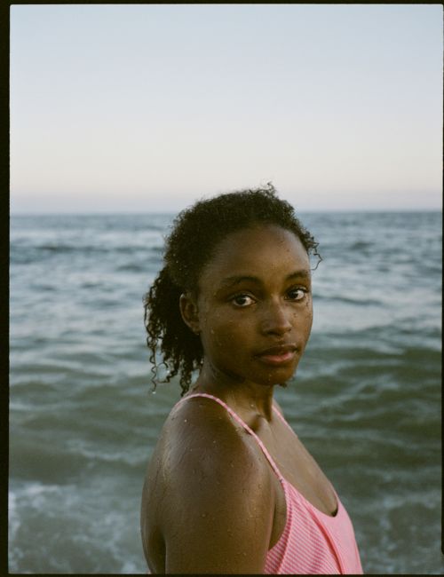 A young woman with dark curly hair stands on a beach, wearing a pink striped swimsuit, with ocean waves and sky in the background.