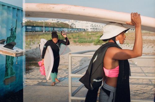Two women at the beach, one carrying a surfboard on her head, the other with a surfboard under her arm near a mural of the Statue of Liberty.