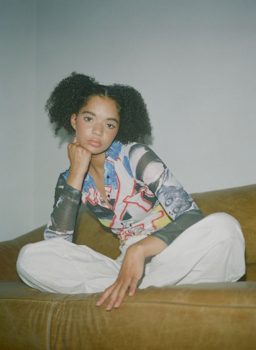 Young woman with afro hairstyle poses on a brown couch in a brightly lit room. She wears a graphic print shirt and light-colored pants.
