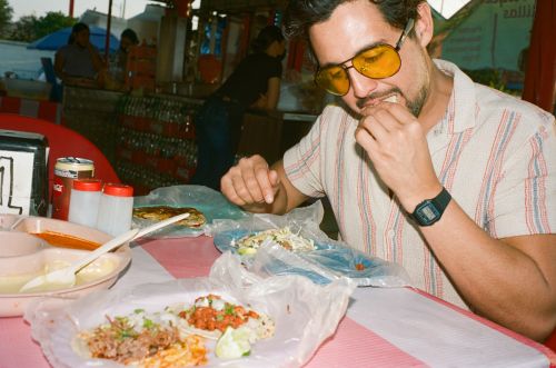 Man in yellow glasses eats tacos at an outdoor stand, wearing a striped shirt with various tacos, bowls, and sauces in the foreground.