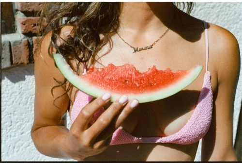 Young adult female in pink bikini top holds partially eaten watermelon slice against white textured wall and brick background in outdoor setting.