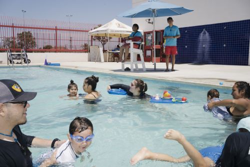 Outdoor swimming pool scene with adults and children enjoying a sunny day, overseen by lifeguards, featuring blue water and a relaxed atmosphere.