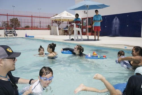 Outdoor swimming pool scene with adults and children enjoying a sunny day, overseen by lifeguards, featuring blue water and a relaxed atmosphere.