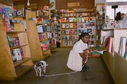 Young woman crouches reading a book while holding the leash of her dog in a brightly lit bookstore with shelves of books.