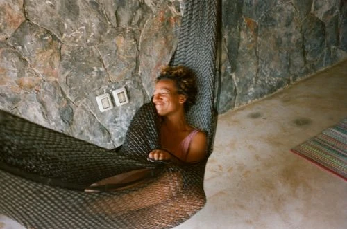 A smiling adult female with curly hair relaxing in a hammock indoors, in front of a stone wall with neutral color tones and natural light.