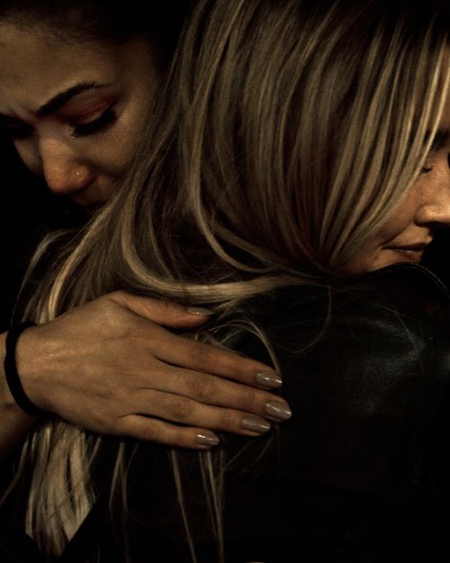 Two adult women embrace tightly against a dark background. One with light blonde hair faces away, and the other with dark hair faces forward.