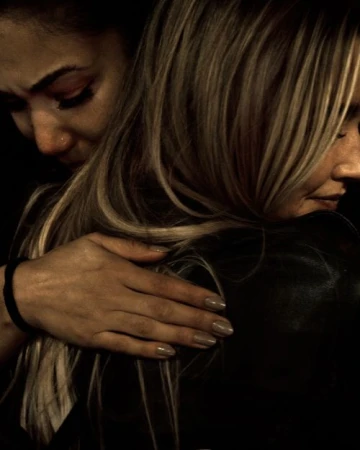 Two adult women embrace tightly against a dark background. One with light blonde hair faces away, and the other with dark hair faces forward.