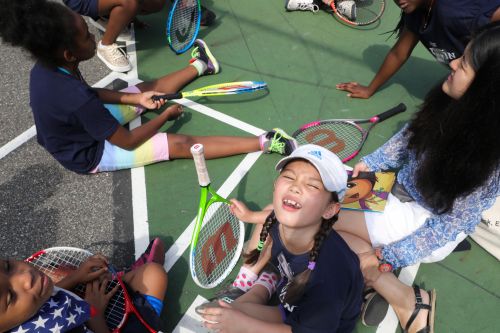 A group of children sitting on a green tennis court surrounded by tennis rackets in the daylight, with sunlight visible on the surface.