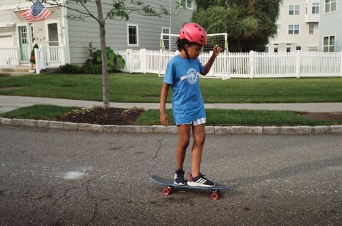 A young child wearing a pink helmet rides a skateboard in a residential area with houses, lawns, and a white picket fence visible.