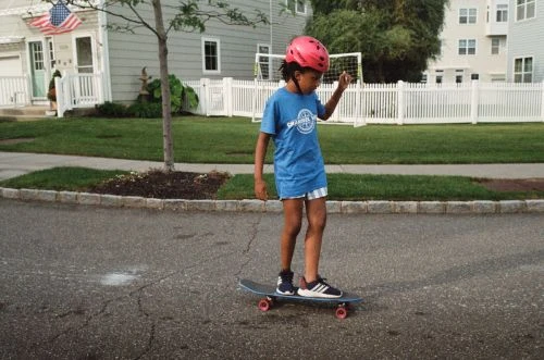 A young child wearing a pink helmet rides a skateboard in a residential area with houses, lawns, and a white picket fence visible.