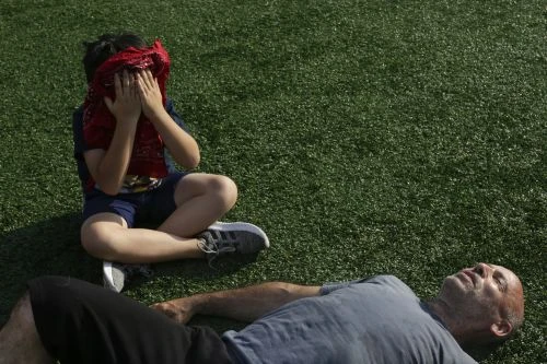 Boy covers his eyes with a red bandana while sitting on the grass next to a man lying on his back looking up at the sky.