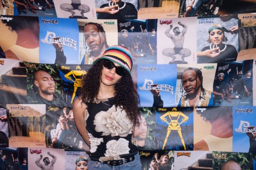 A woman with curly hair and sunglasses poses in front of a wall covered in magazine covers, wearing a floral top and a colorful hat.