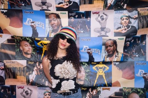 A woman with curly hair and sunglasses poses in front of a wall covered in magazine covers, wearing a floral top and a colorful hat.
