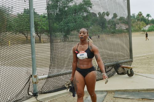 Female athlete standing outdoors on a track, post competition, wearing athletic wear, with a discus cage and trees in the background.
