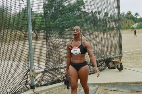 Female athlete standing outdoors on a track, post competition, wearing athletic wear, with a discus cage and trees in the background.
