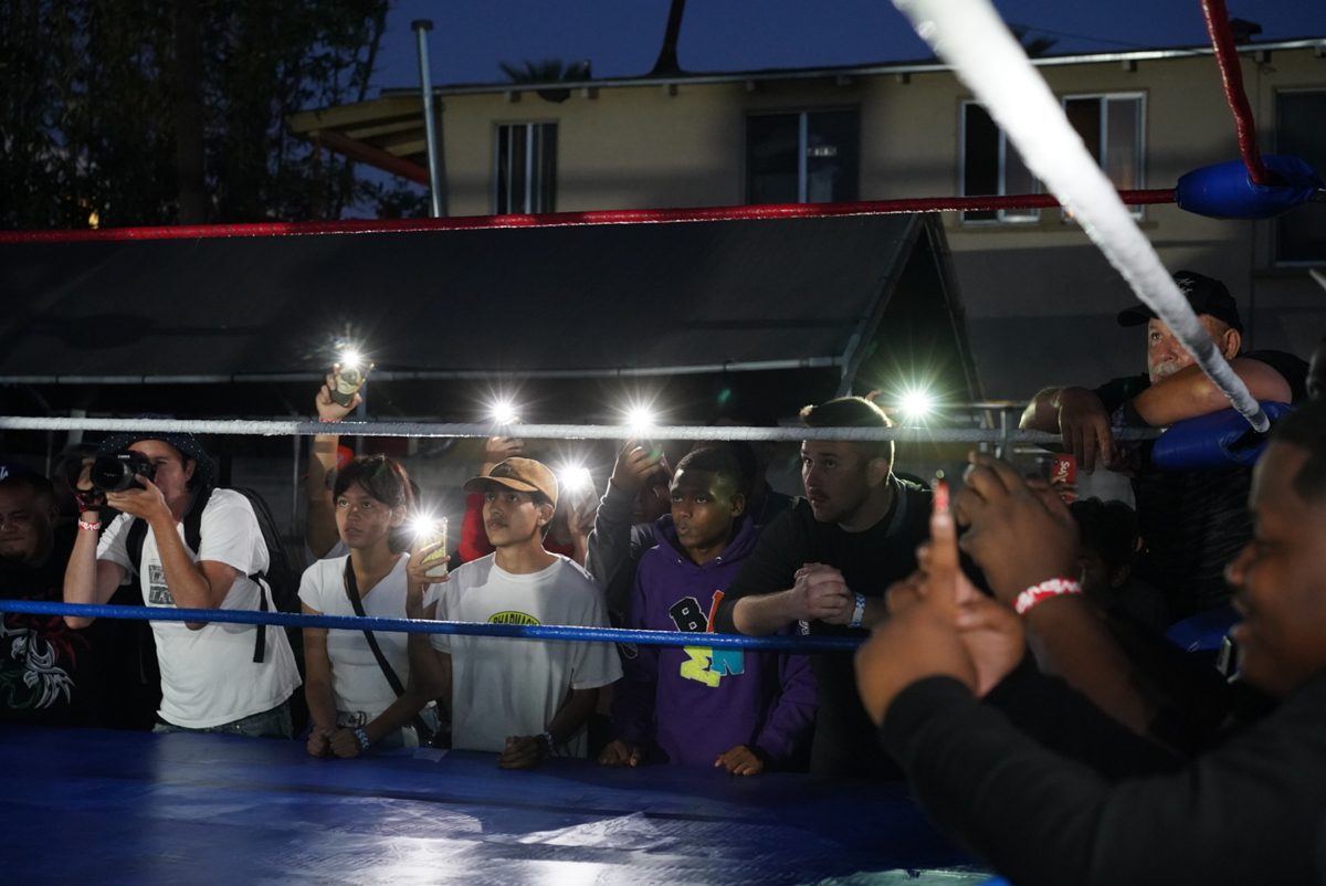 Crowd watches wrestling match, cell phone lights illuminate the scene. People gather around the wrestling ring in an outdoor setting.
