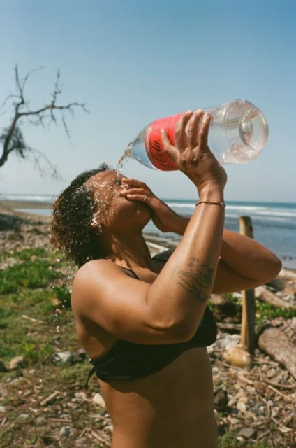 Woman in bikini outdoors pouring water from plastic bottle onto face for refreshment at the beach on sunny day.