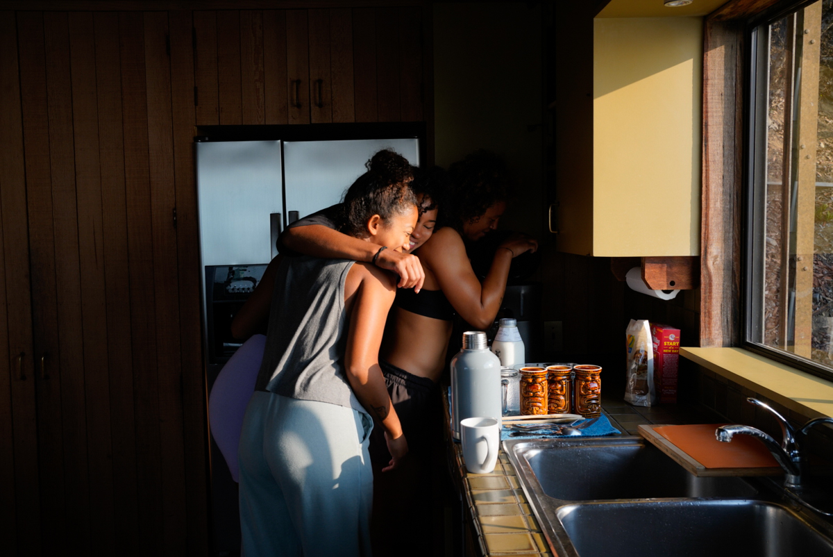 Three women embracing in a sunlit kitchen with stainless steel appliances, mugs, and a window offering a glimpse of the outdoors.