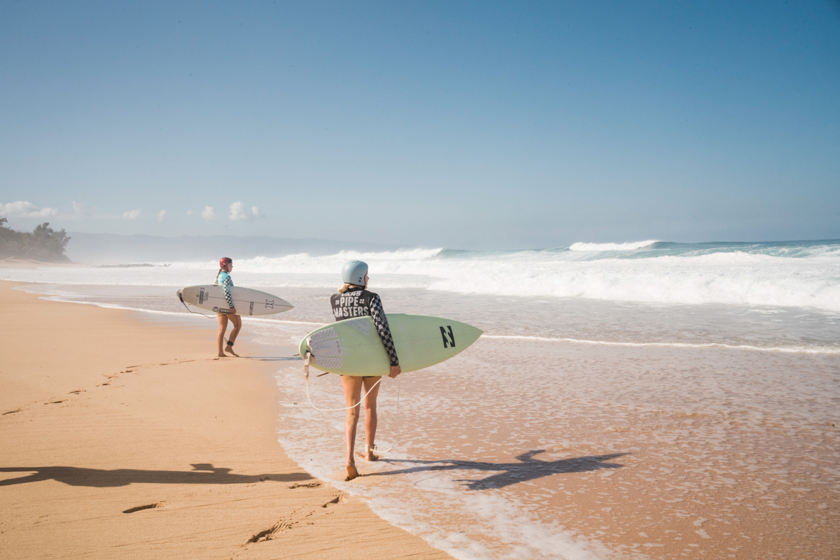 Two women surfers walk along a sunny beach with surfboards. Ocean waves break in the background under a clear sky.
