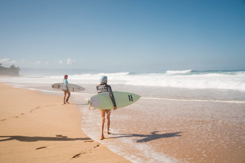 Two women surfers walk along a sunny beach with surfboards. Ocean waves break in the background under a clear sky. 
