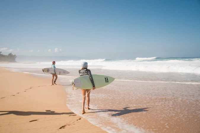 Two women surfers walk along a sunny beach with surfboards. Ocean waves break in the background under a clear sky. 