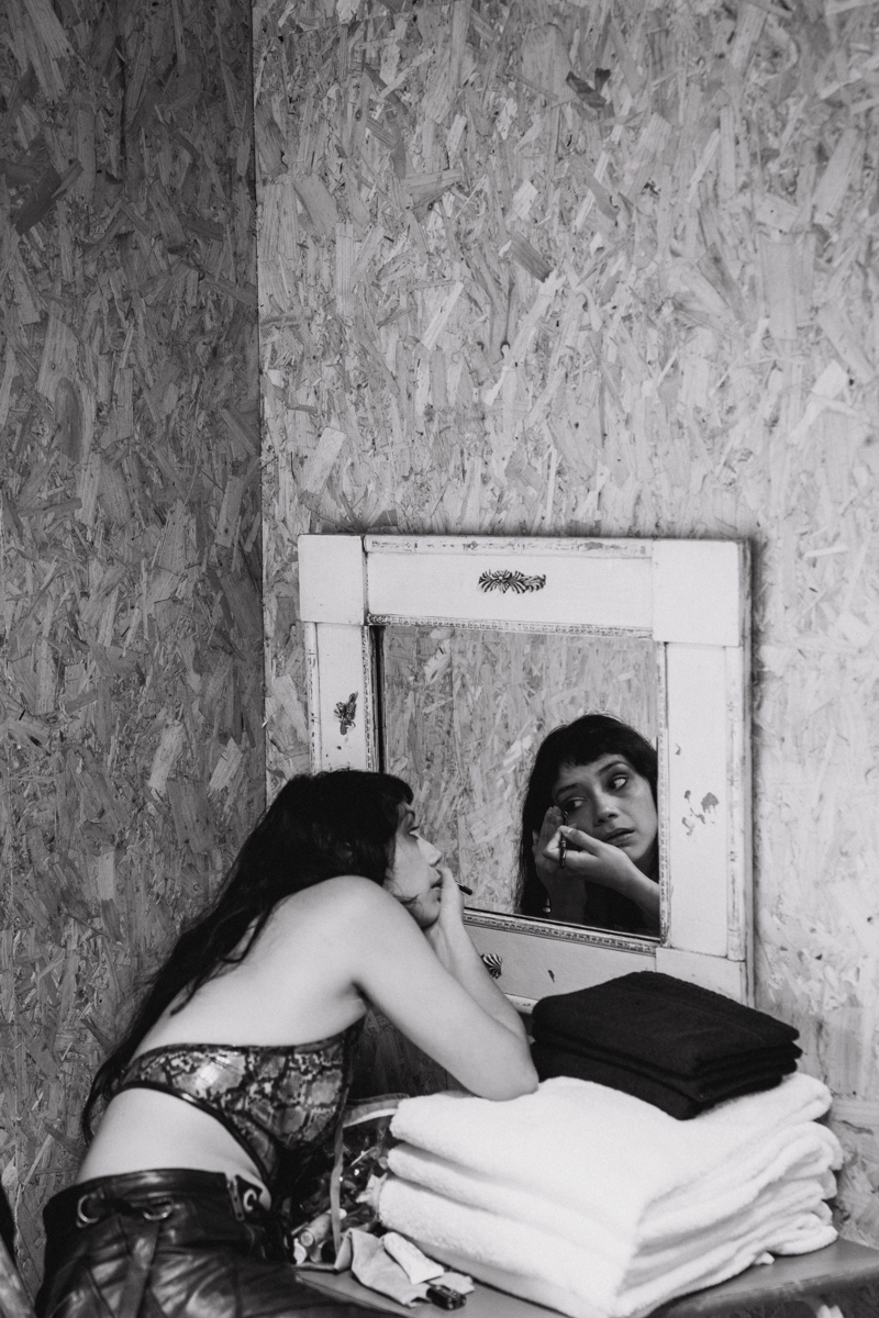Woman applies makeup in an indoor setting near a mirror, white towels, and plywood wall. Black and white photograph shows beauty routine.