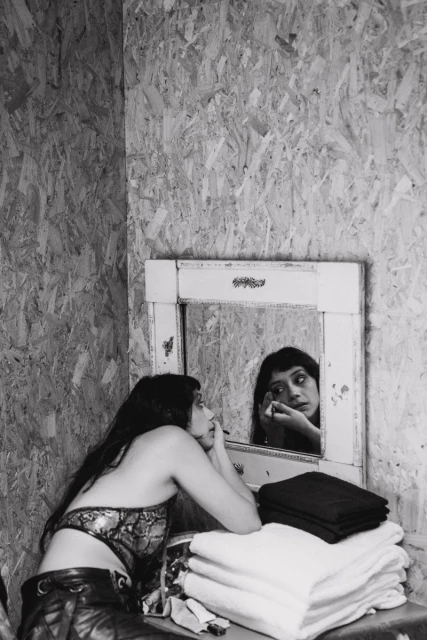 Woman applies makeup in an indoor setting near a mirror, white towels, and plywood wall. Black and white photograph shows beauty routine.