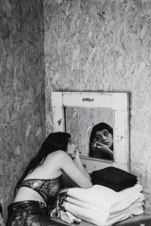 Woman applies makeup in an indoor setting near a mirror, white towels, and plywood wall. Black and white photograph shows beauty routine.
