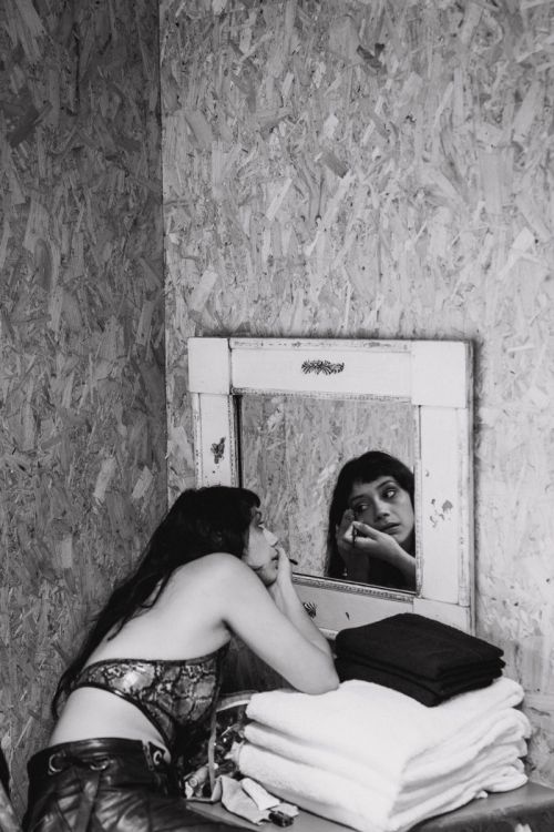 Monochrome photo of a woman applying makeup in a room with textured wooden walls, a mirror, and stacked white and black towels.