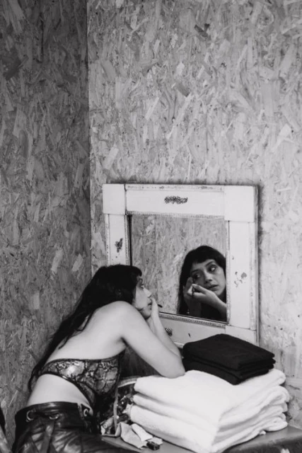 Monochrome photo of a woman applying makeup in a room with textured wooden walls, a mirror, and stacked white and black towels.