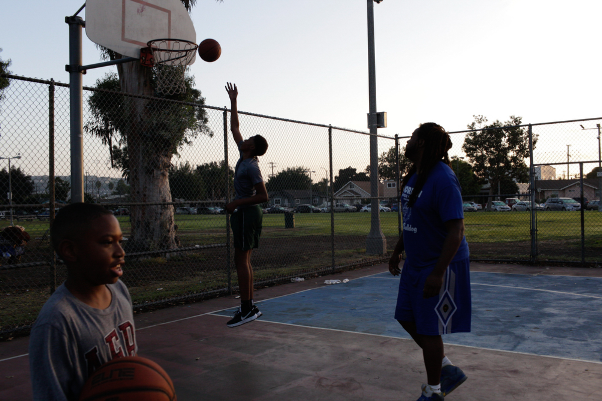 A group of people are playing basketball outside on a concrete court with chain link fences and trees in the background during the day.