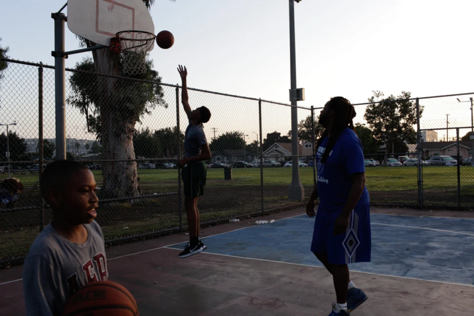 A group of people are playing basketball outside on a concrete court with chain link fences and trees in the background during the day.