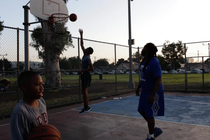 A group of people are playing basketball outside on a concrete court with chain link fences and trees in the background during the day.