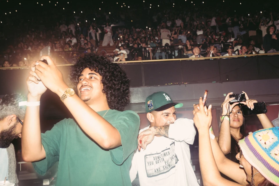 A crowd of young adults at a concert venue, many holding up phones to record the performance, illuminated by the venue lights.