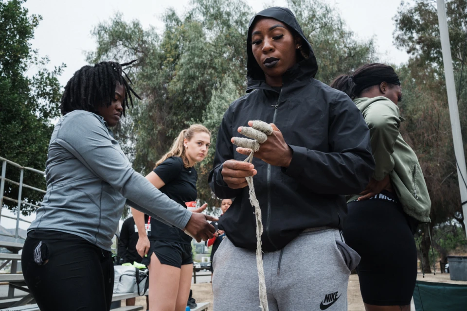 Female athlete wraps her hands with athletic tape, other athletes stand in the background in an outdoor training setting.