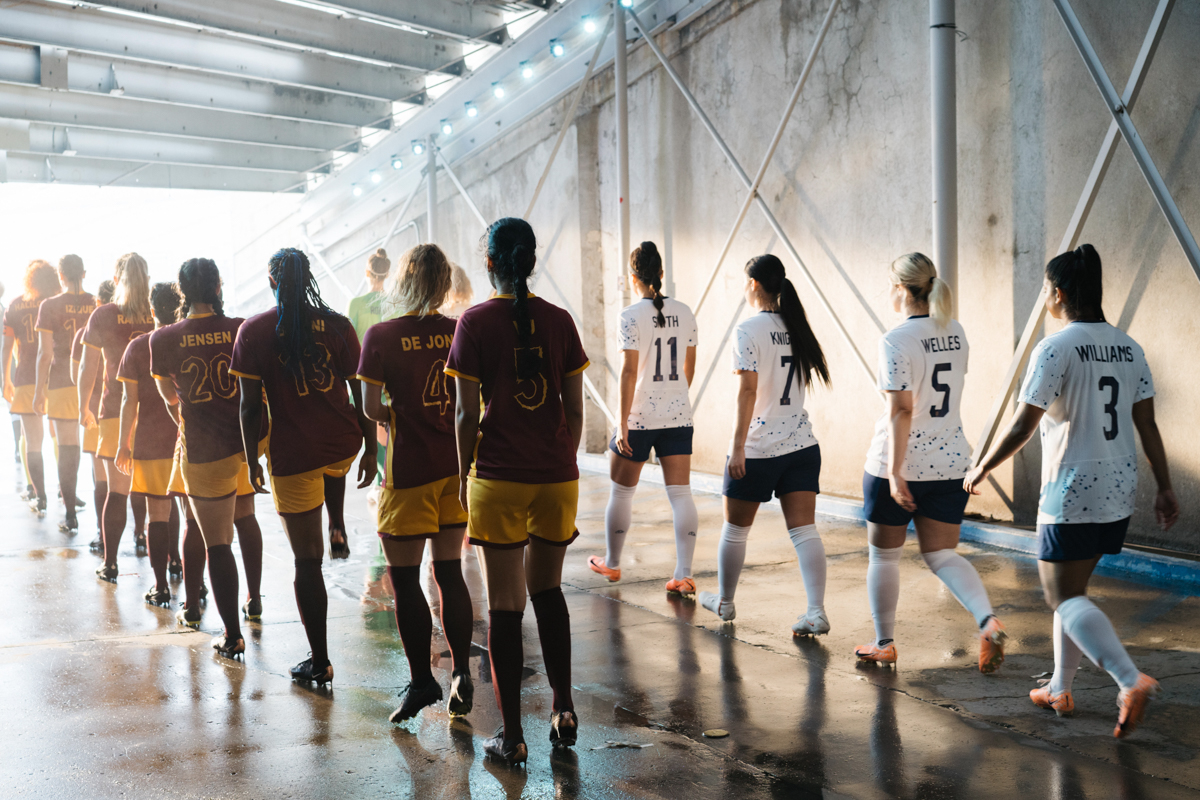 Female soccer players in maroon and white uniforms walk through tunnel with bright lighting and concrete walls to play a game of soccer.
