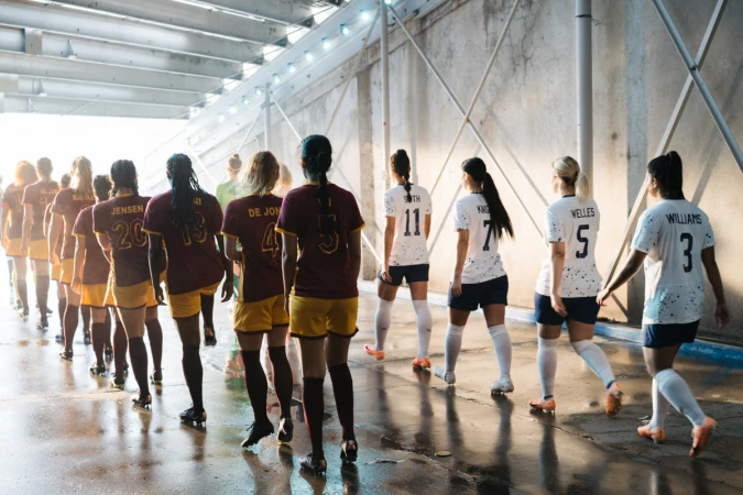 Female soccer players in maroon and white uniforms walk through tunnel with bright lighting and concrete walls to play a game of soccer.